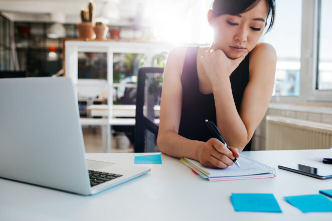 Shot of asian businesswoman writing notes on notepad. Female executive sitting at her desk with laptop taking notes.