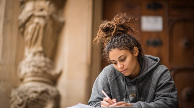 Student writing notes while sitting outdoors near a historic building